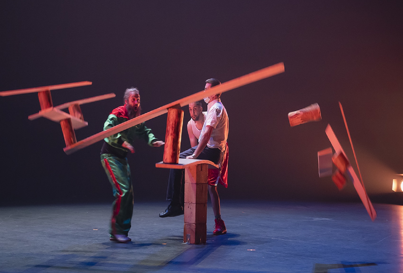 Tres personas en escena. Una de ellas está sentada en una tabla, elevada por una columna de ladrillos, que hace de balanza y en el otro lado hay unos troncos para equilibrar. Las otras dos personas están de pie y miran como se caen los troncos de madera que equilibraban la balanza.